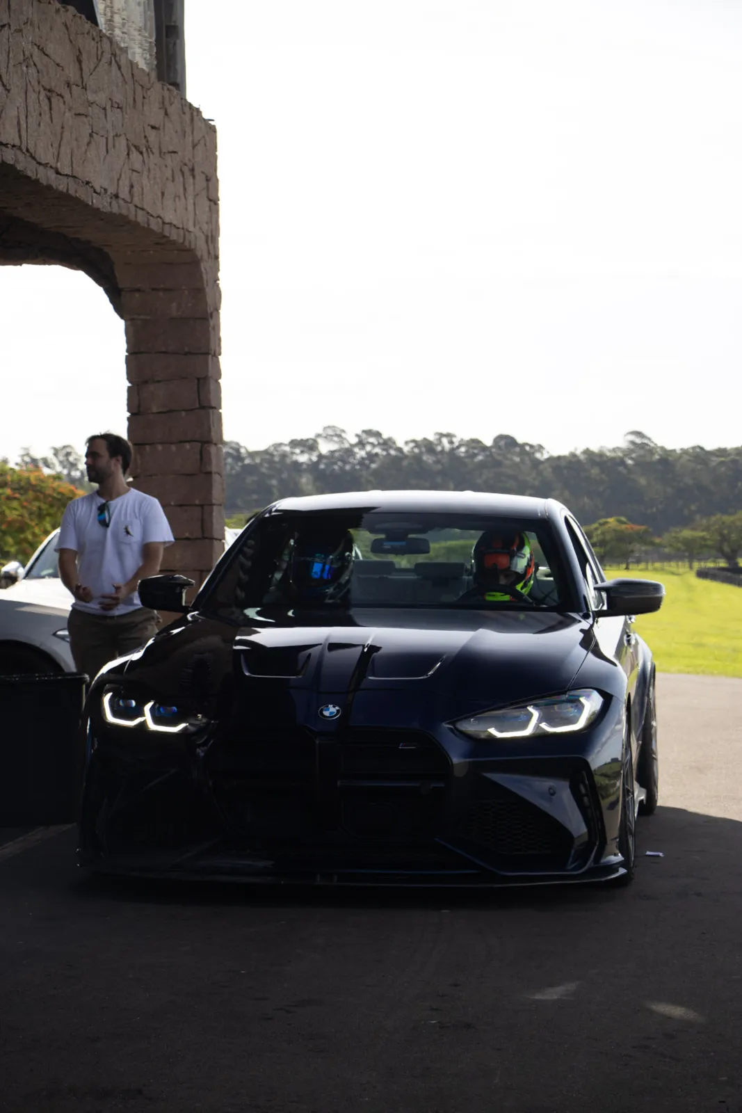 BMW M3 preto com pilotos de capacete no paddock